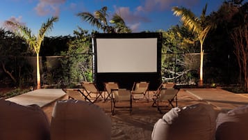 Un écran de cinéma en plein air, sur lequel rien n’est projeté pour le moment, entouré de fauteuils et de poufs, avec en arrière-plan des palmiers et un ciel dégagé.