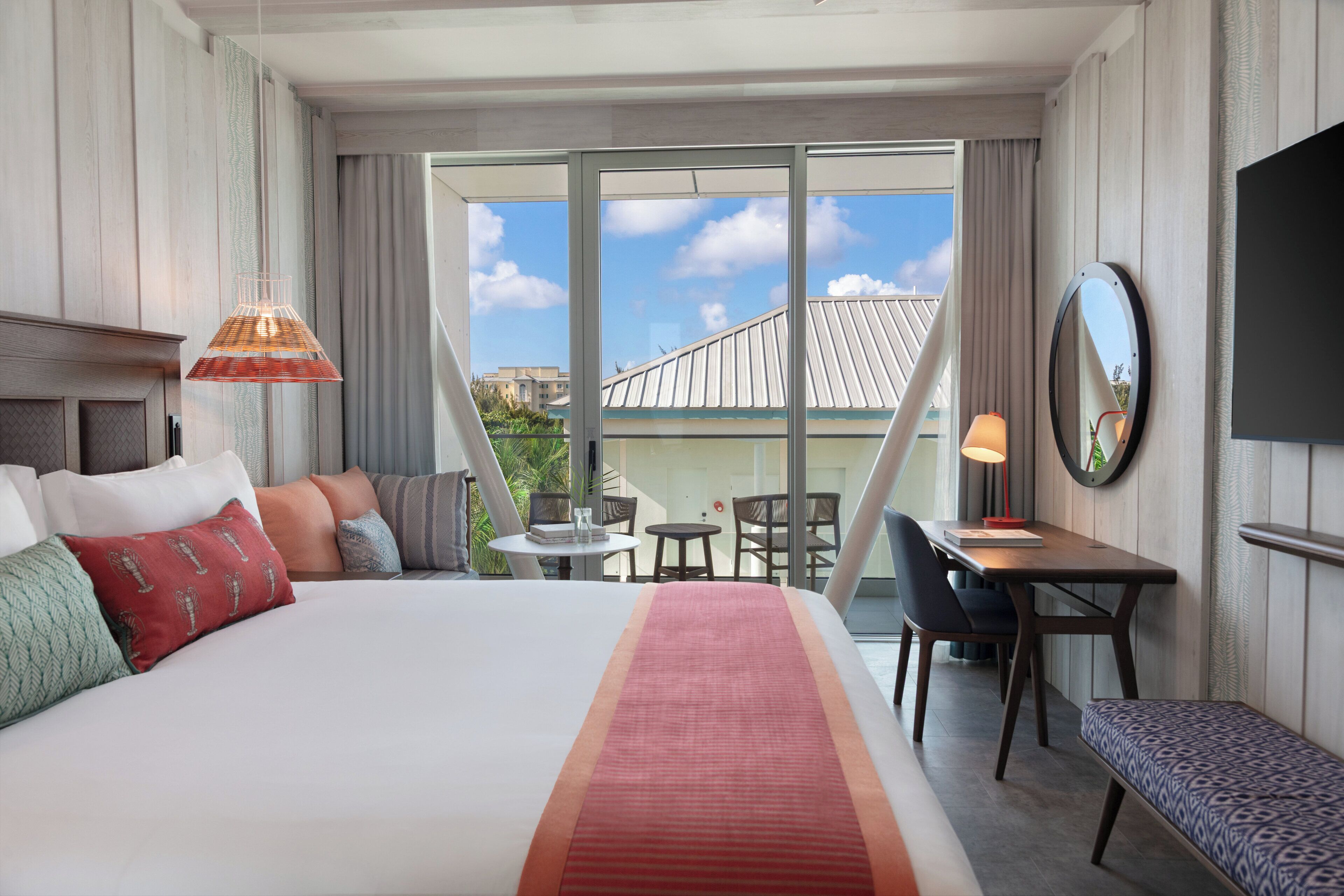 A hotel room with a bed, a desk, a chair, a table, a bench, a TV, and a large window with a view of buildings.