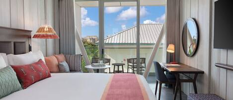 A hotel room with a bed, a desk, a chair, a table, a bench, a TV, and a large window with a view of buildings.