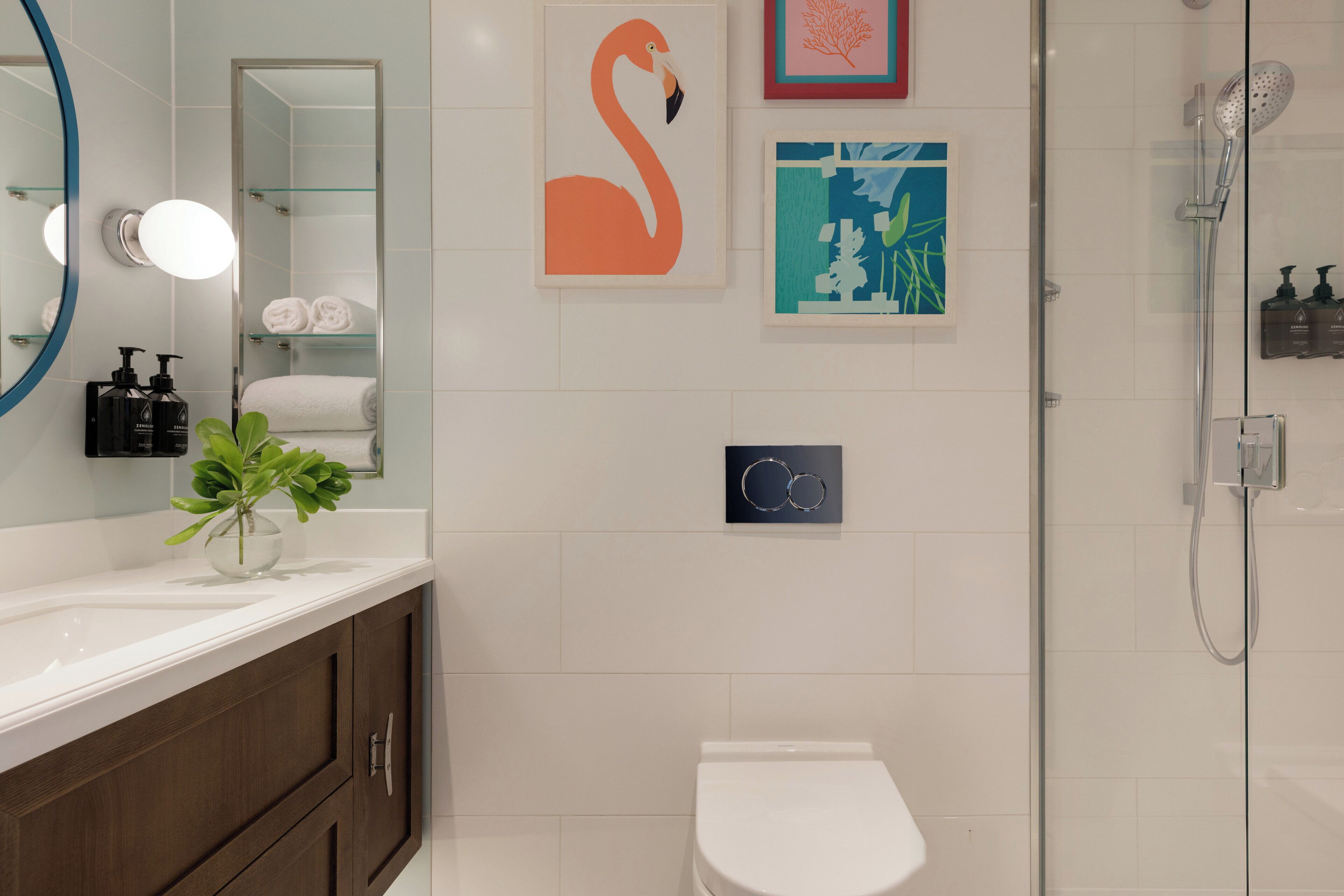 Et moderne badeværelse med toilet i hvid, glasvæg omkring brusebad, spejl med lys og kunst på væggene.
