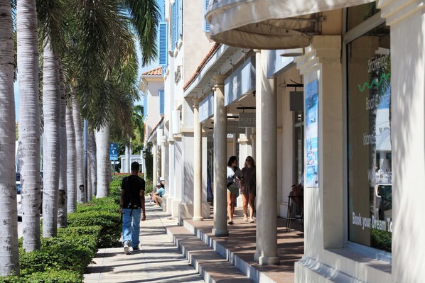 A pedestrian walkway lined with palm trees and buildings with large windows and columns.