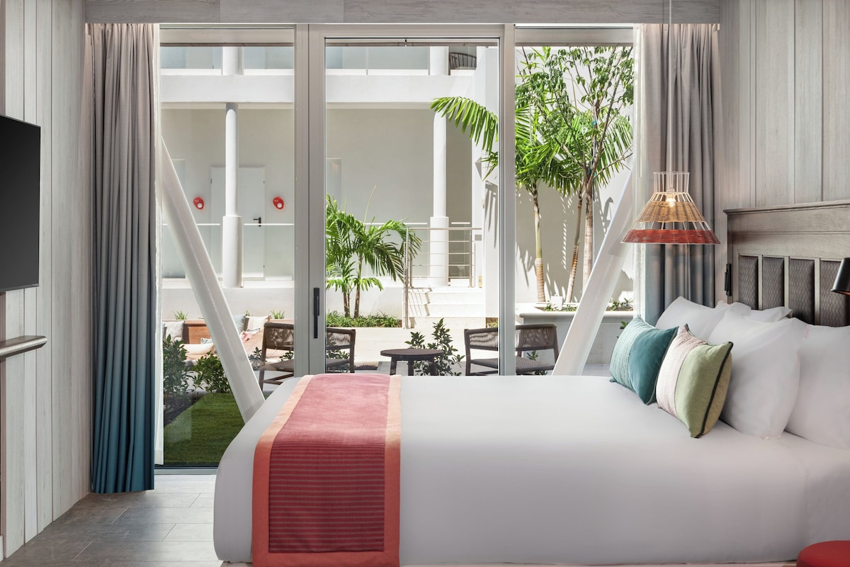 A hotel room with a bed, a TV, a balcony with plants, and a view of the outdoors.