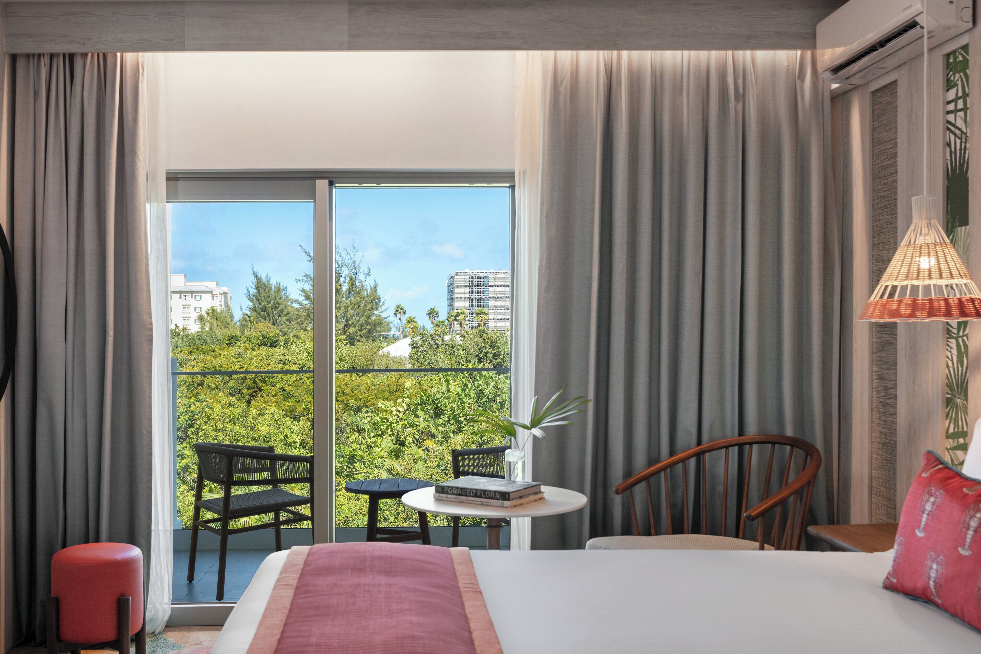 Habitación de hotel con una cama, una pequeña mesa con libros, una silla, un taburete rojo y vistas a zonas verdes y edificios a través de un ventanal grande.