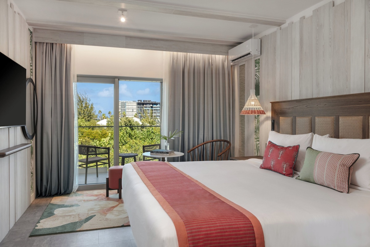 A hotel room with a large bed, a TV, a balcony with a table and chairs, and a view of buildings and greenery.