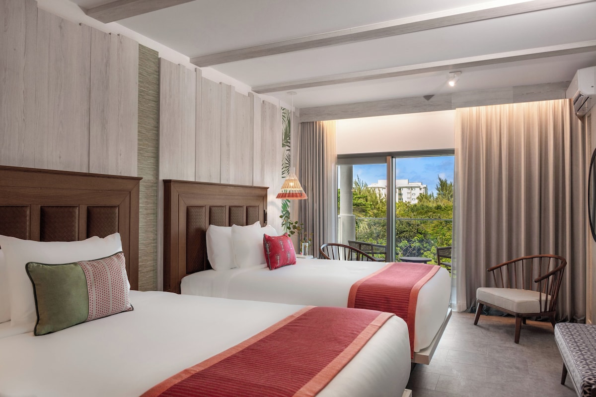 A hotel room with two beds, a balcony with a view, and a seating area with a chair and a bench.
