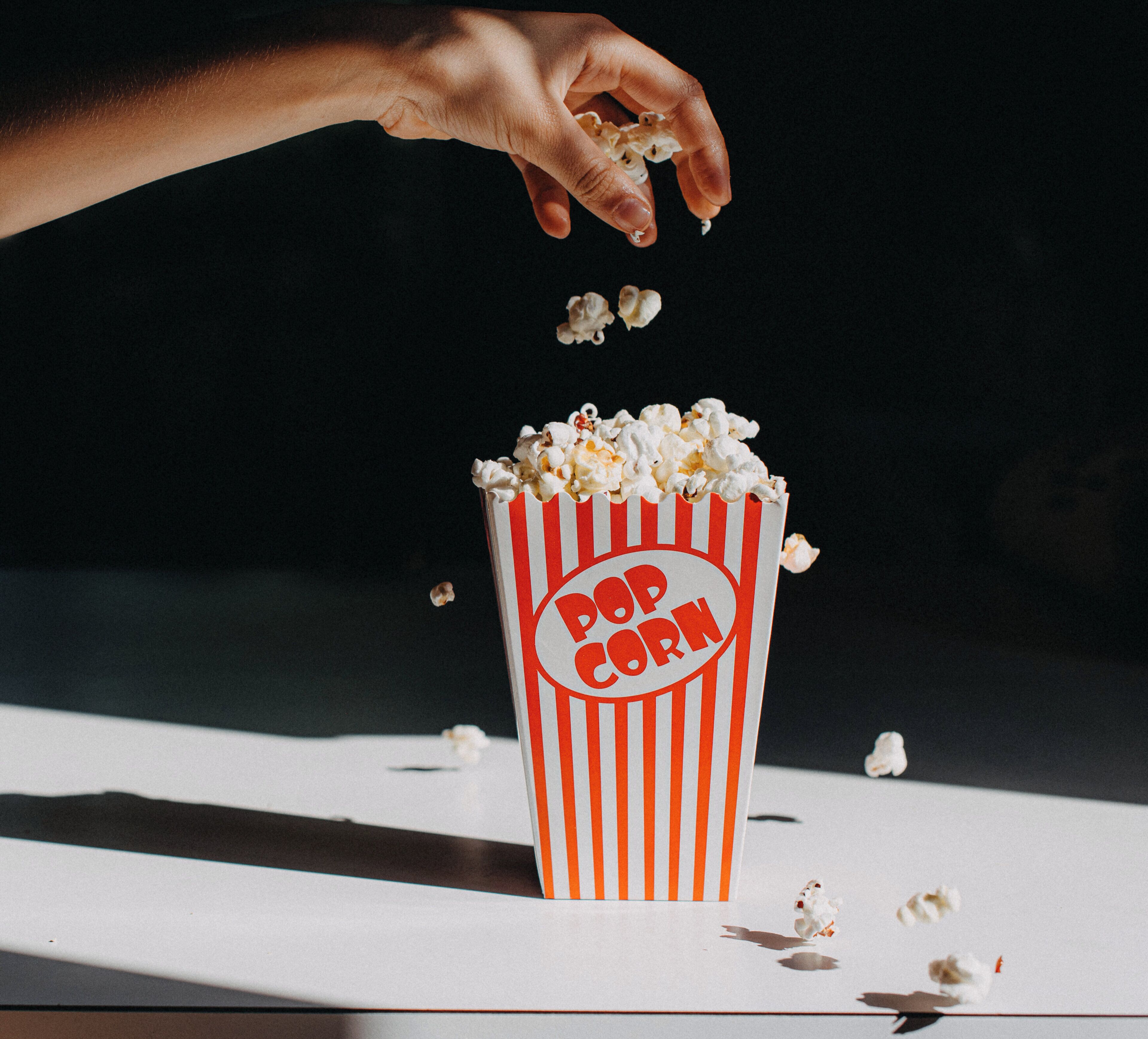 Eine Hand lässt Popcorn in einen gestreiften Popcornbehälter fallen.
