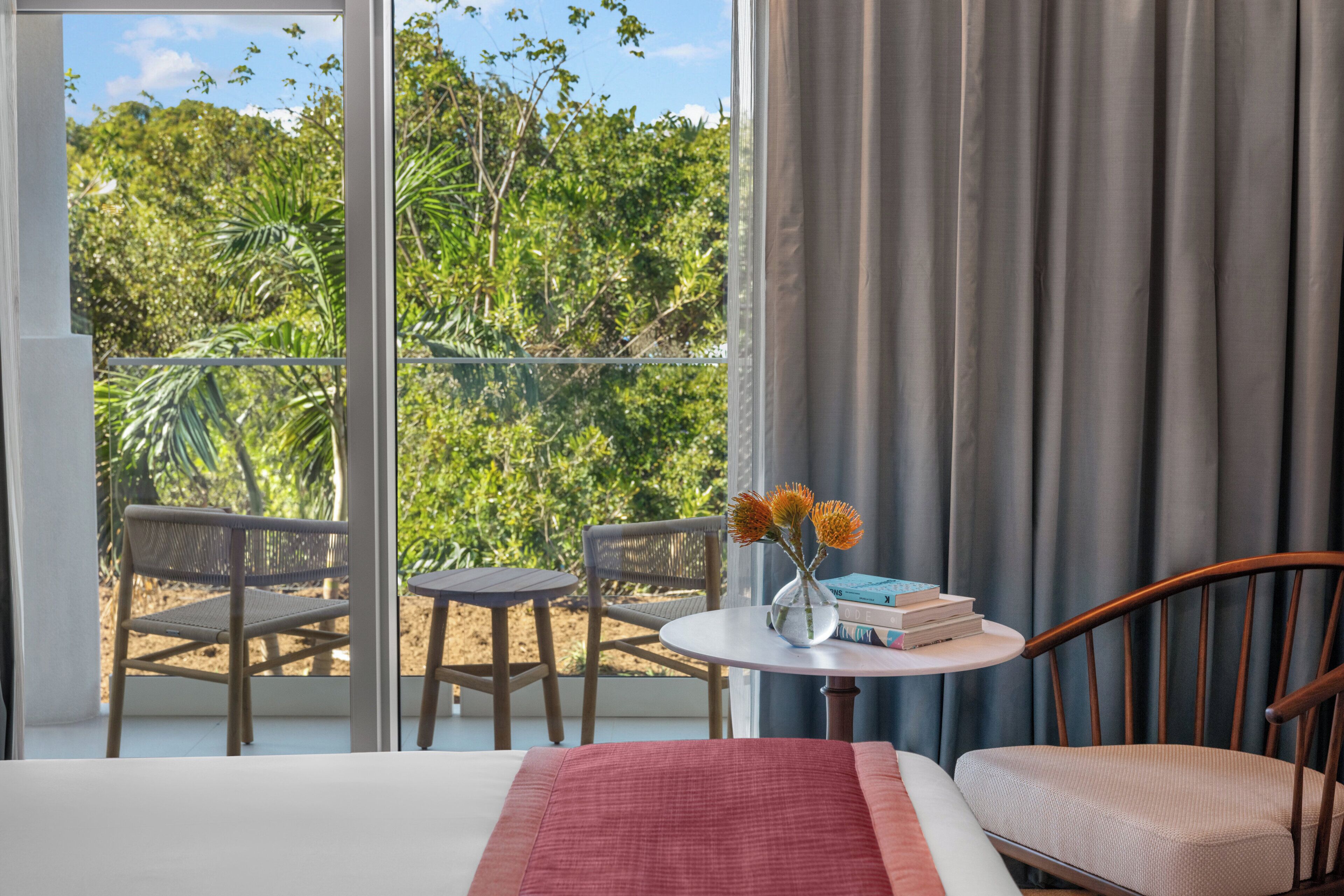 A hotel room with a bed, a small table with a vase of flowers, two chairs, and a view of greenery through a large window.