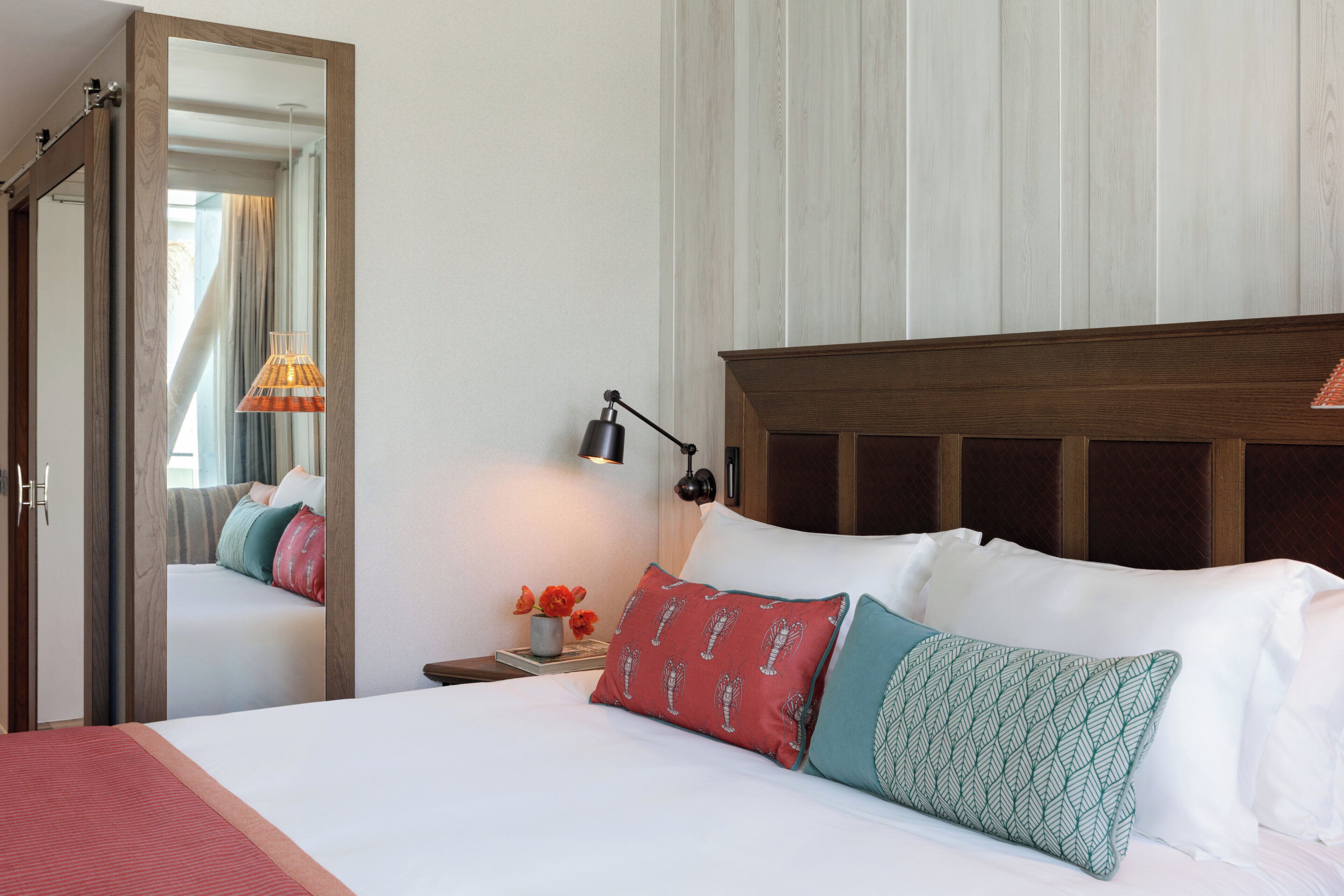 A hotel room with a wooden headboard, a bed with white linens, a nightstand with a lamp, and a mirror reflecting part of the room.