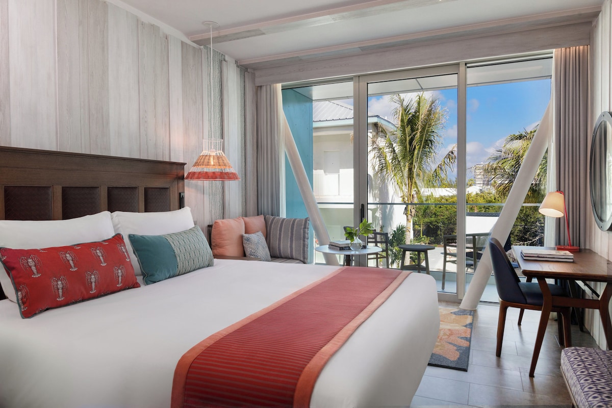 A hotel room with a large bed, a desk, and a balcony with a view of palm trees.