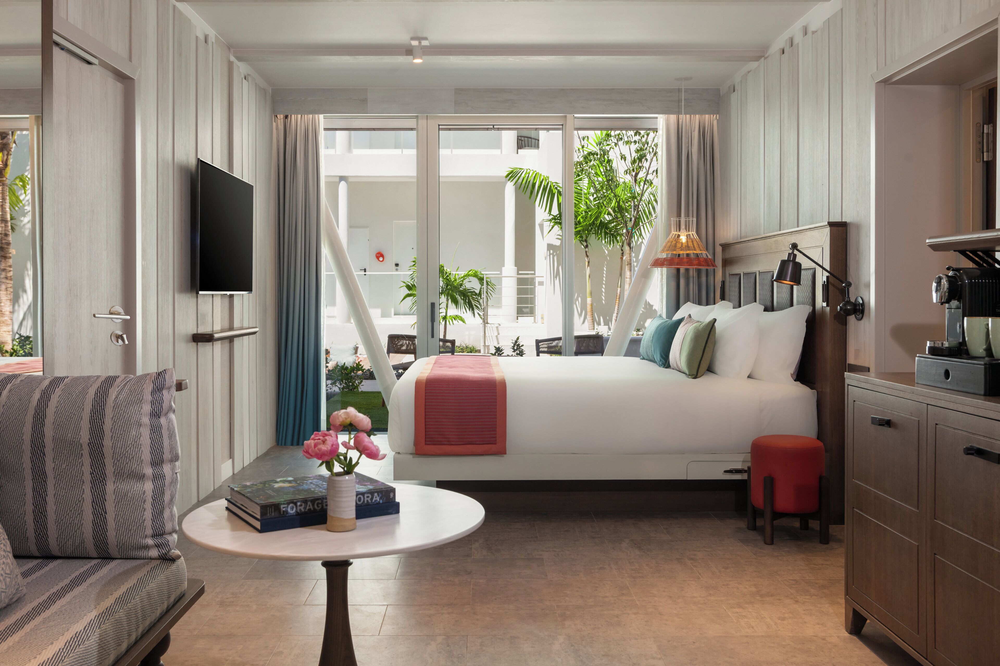 Quarto com cama, televisão, mesa com livros e flores, um banquinho vermelho e vista para o exterior.