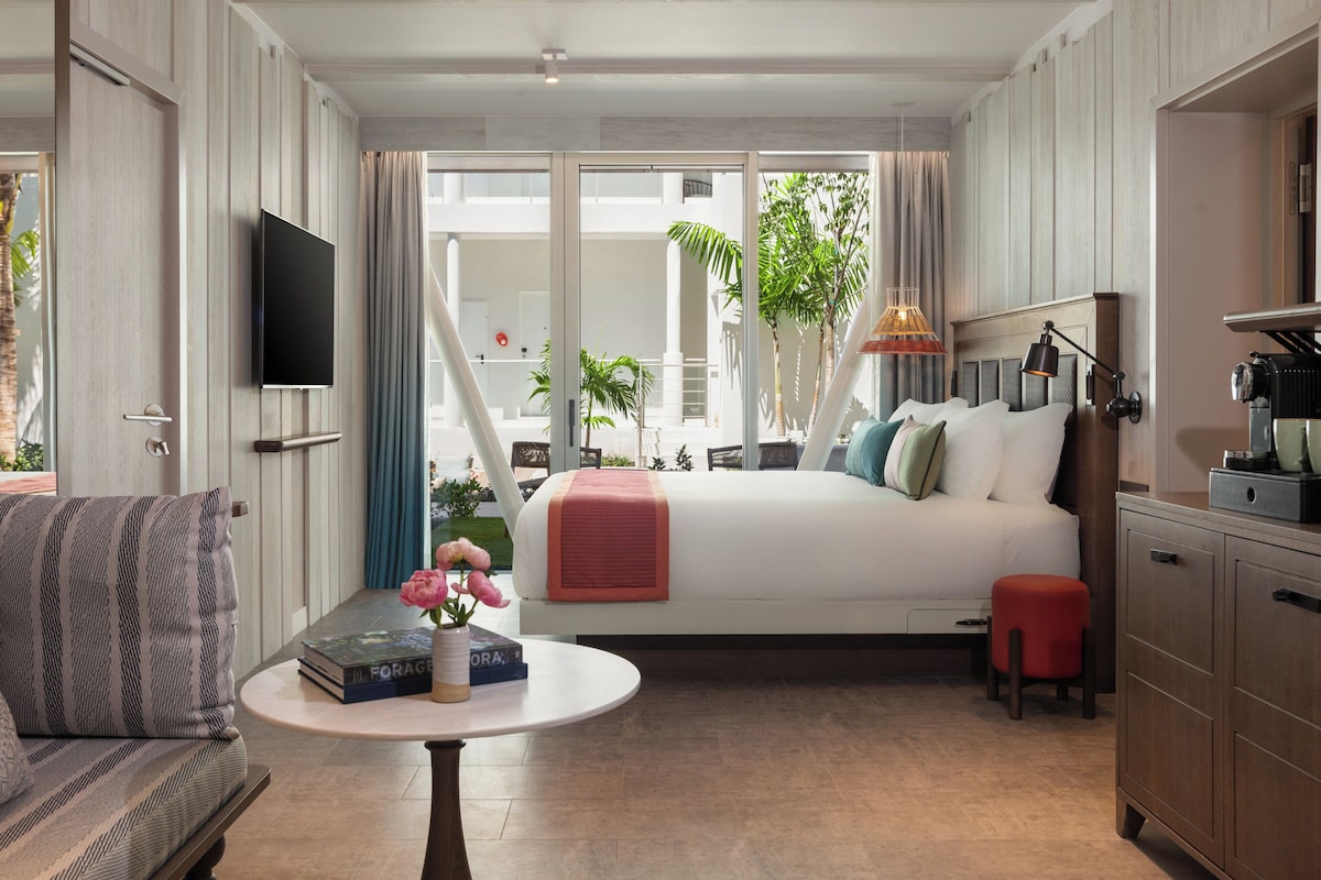 A bedroom with a bed, a TV, a table with books and flowers, a red stool, and a view of the outside.