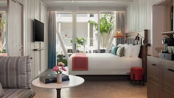 Quarto com cama, televisão, mesa com livros e flores, um banquinho vermelho e vista para o exterior.