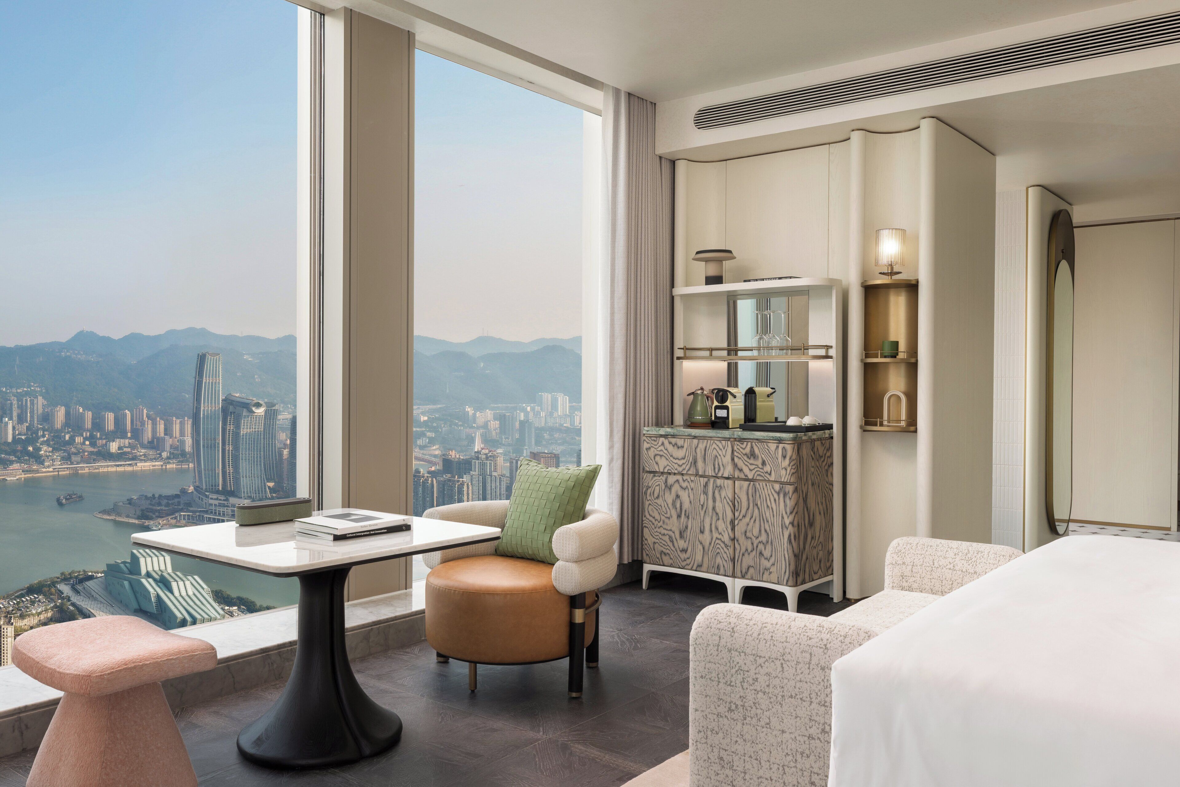 A hotel room with a large window offering a city view, a desk with a chair, a bed, and a minibar.