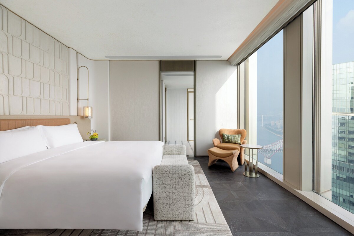 Una habitación de hotel moderna con una cama grande, un área de descanso con una silla y una mesa, y un amplio ventanal con vistas a la ciudad.