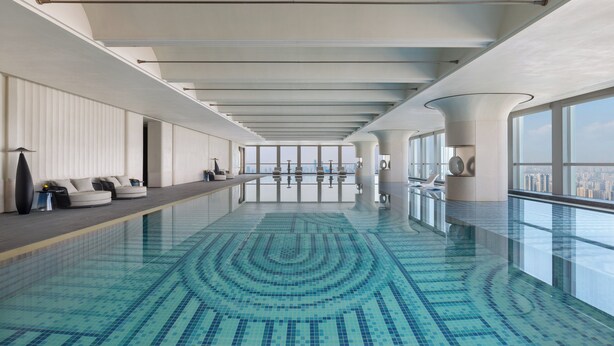 A spacious indoor swimming pool with a tiled floor, a long bench along one side, and large windows offering a city view.