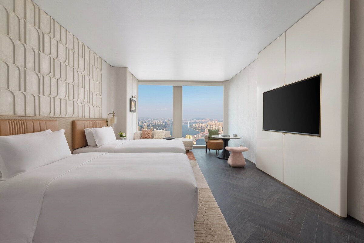 Una habitación de hotel moderna con dos camas, un televisor grande y vistas a un paisaje urbano.