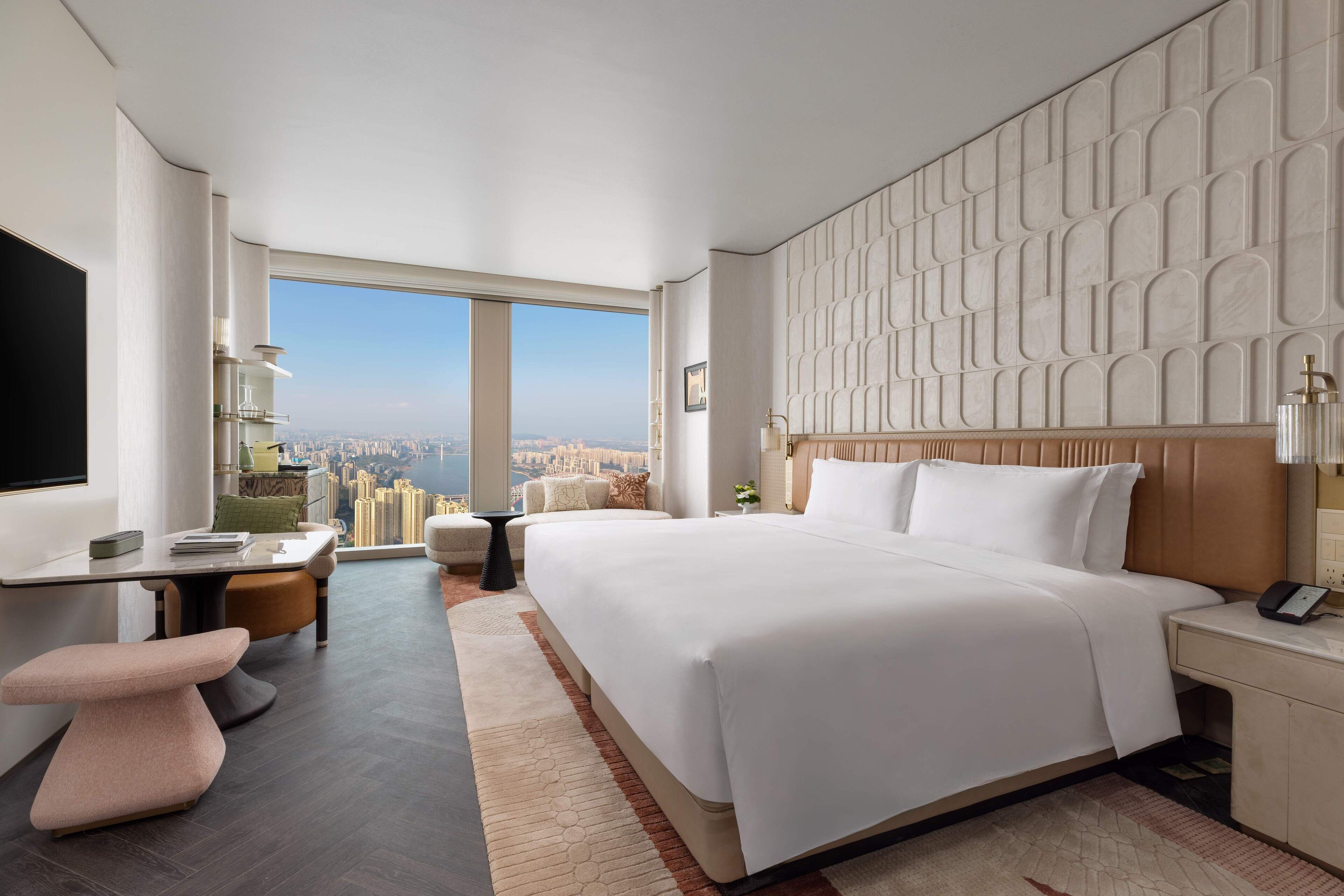 A modern hotel room with a large bed, a desk, a chair, and a view of the city.