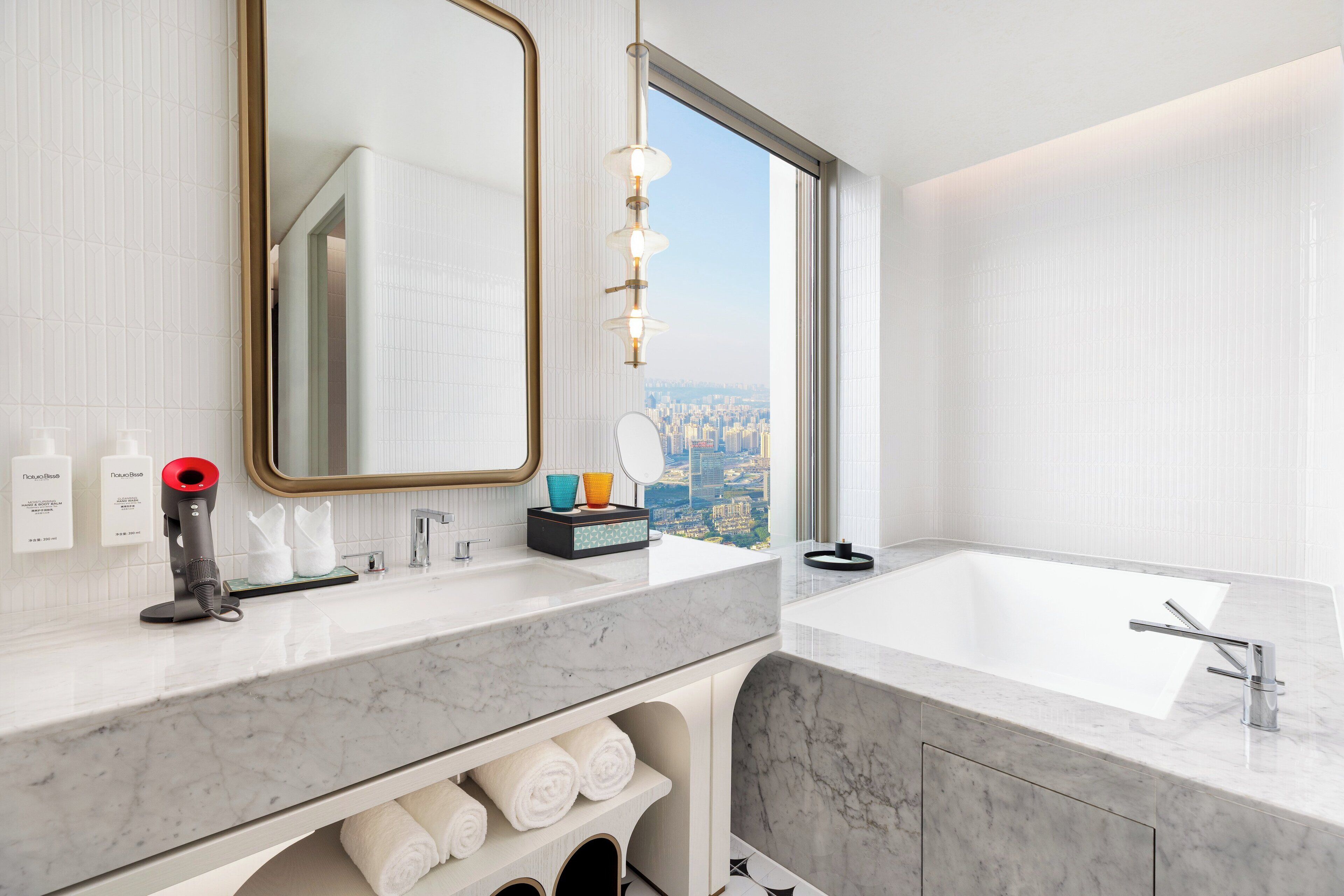 A modern bathroom with a large mirror, a sink, and a bathtub. There is a towel rack with towels, a hairdryer, and a box of toiletries. The bathroom has a large window with a view of a cityscape.