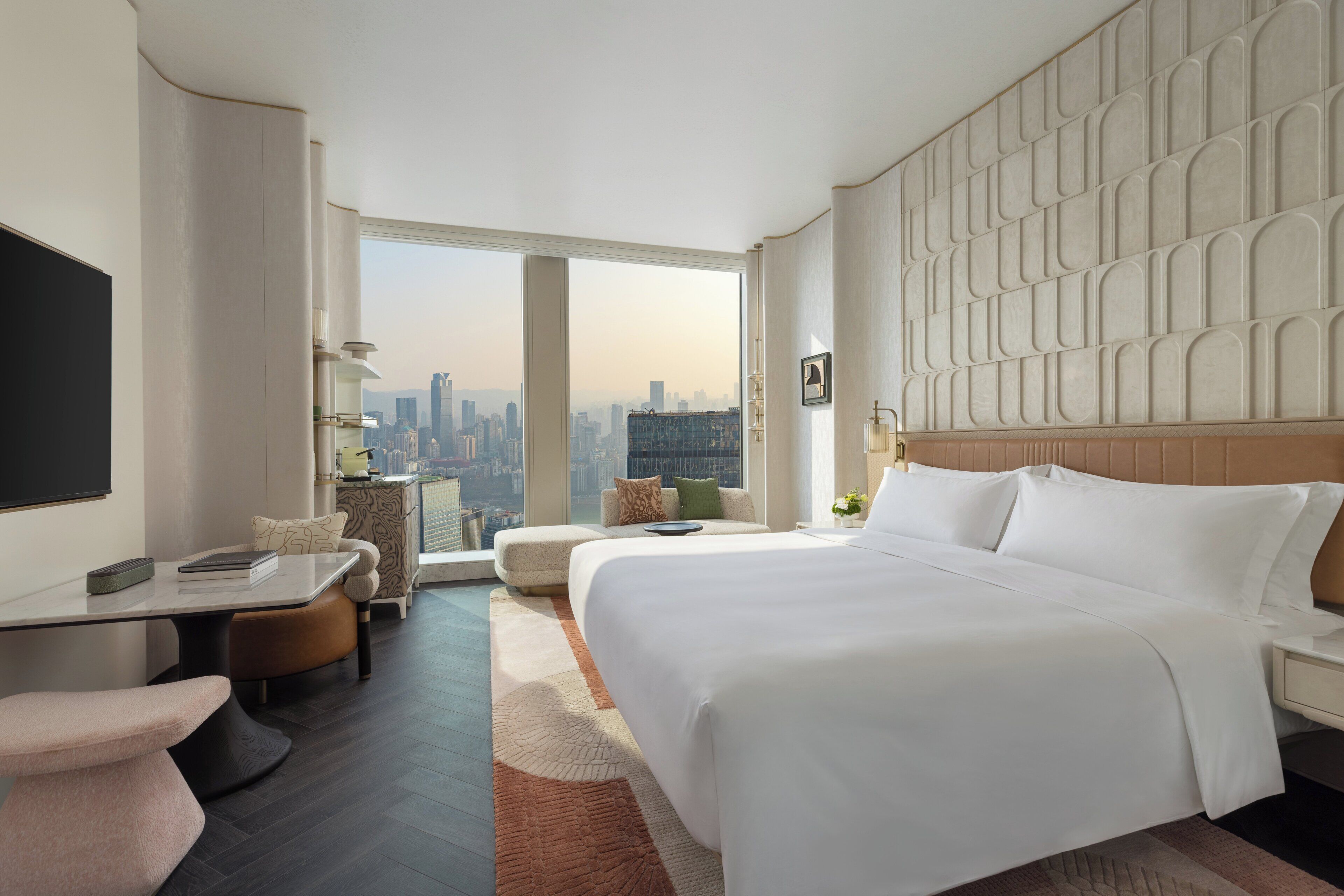Ein modernes Hotelzimmer mit einem großen Bett, einem Schreibtisch, einem Sessel und Blick auf die Stadtlandschaft.