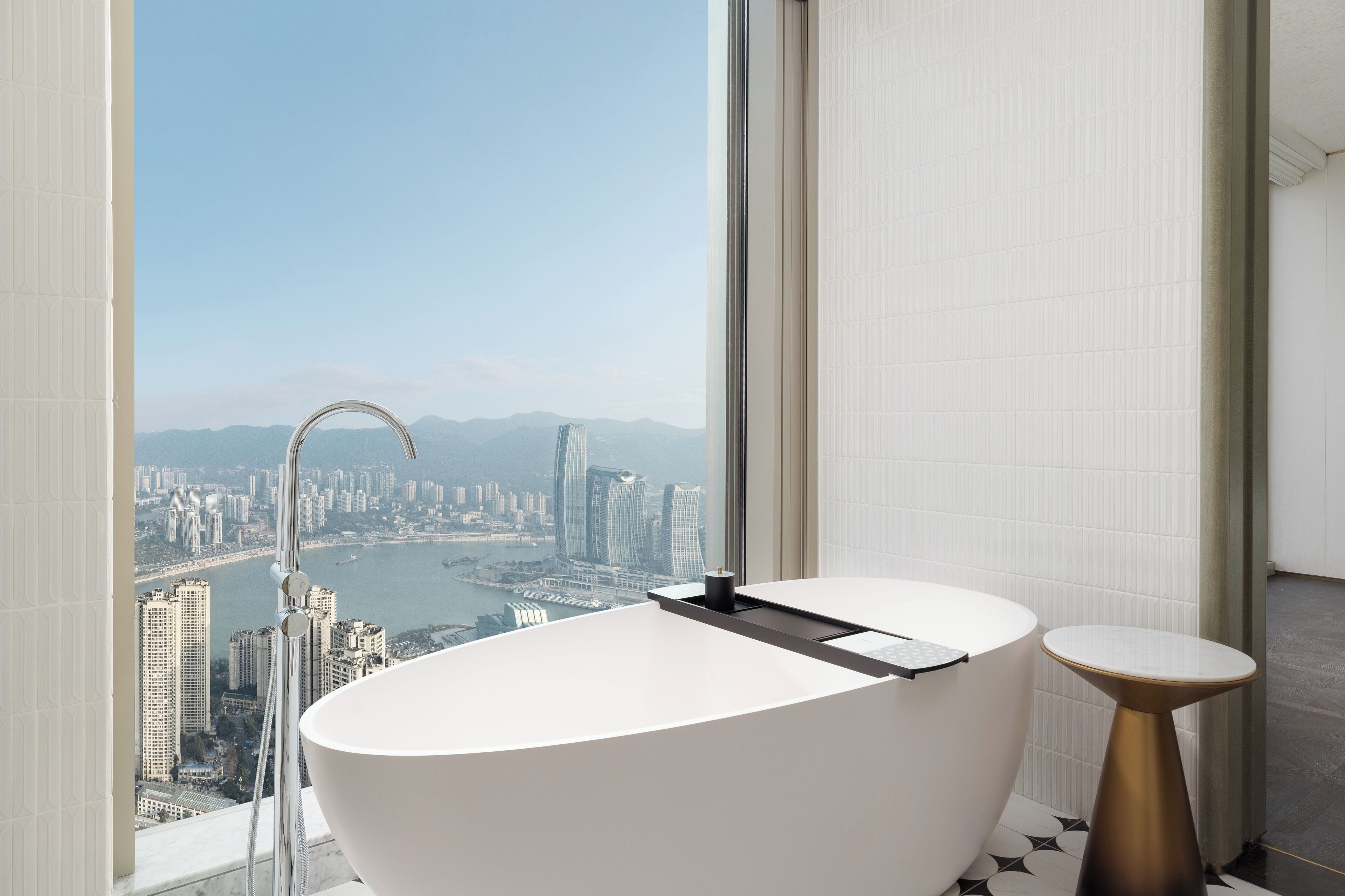 A modern bathroom with a freestanding bathtub, a small round table, and a large window offering a cityscape view.