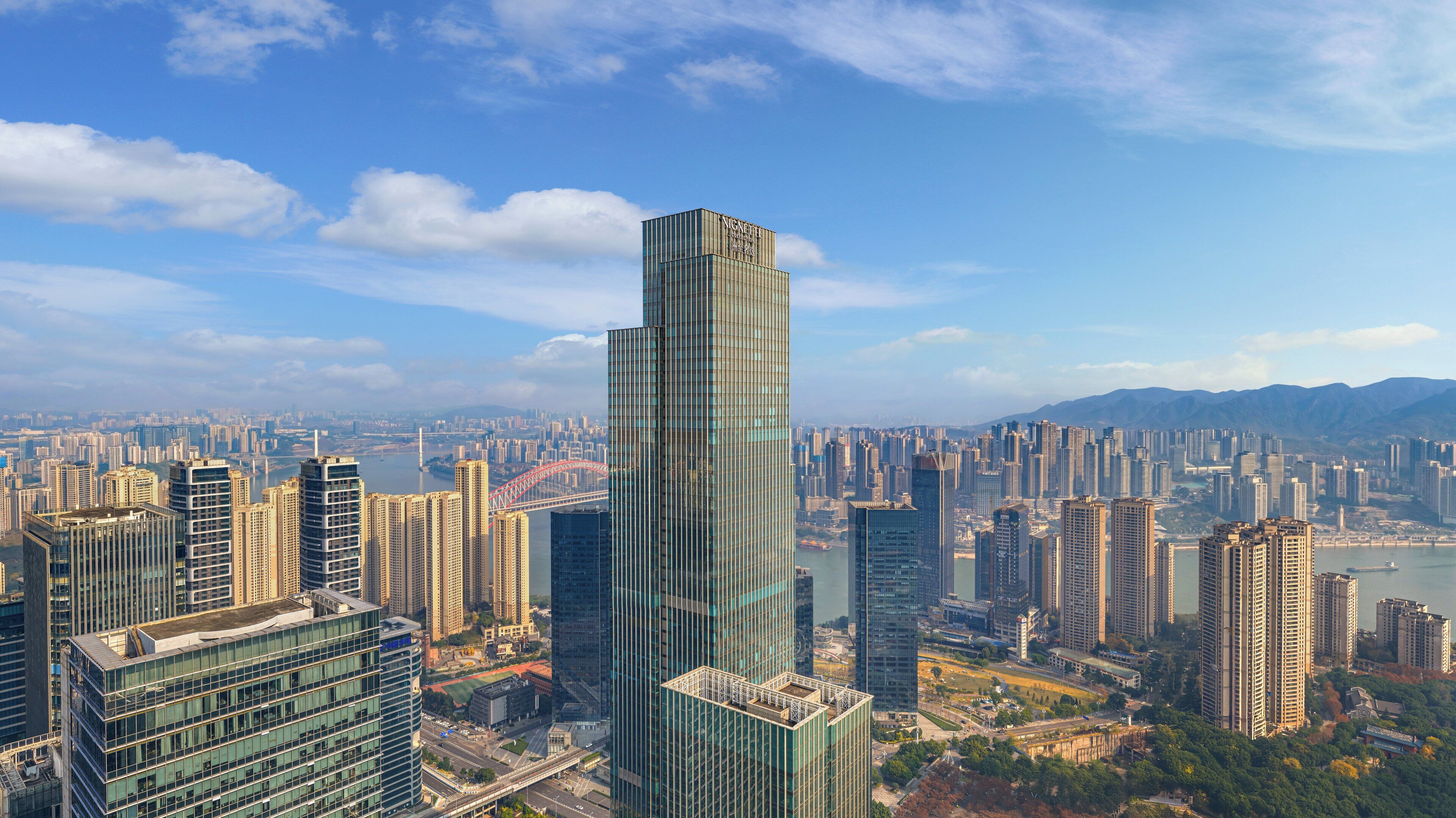 A high-rise building with a modern glass facade, surrounded by a dense urban skyline and a river.