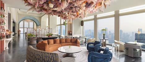 A spacious living area with a large round coffee table, sofas, and chairs. There is a city view through large windows.