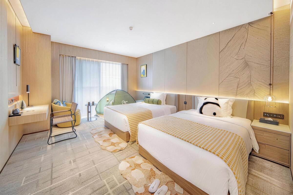 A modern hotel room with two beds, a desk, a chair, a small table, and a large window with curtains.