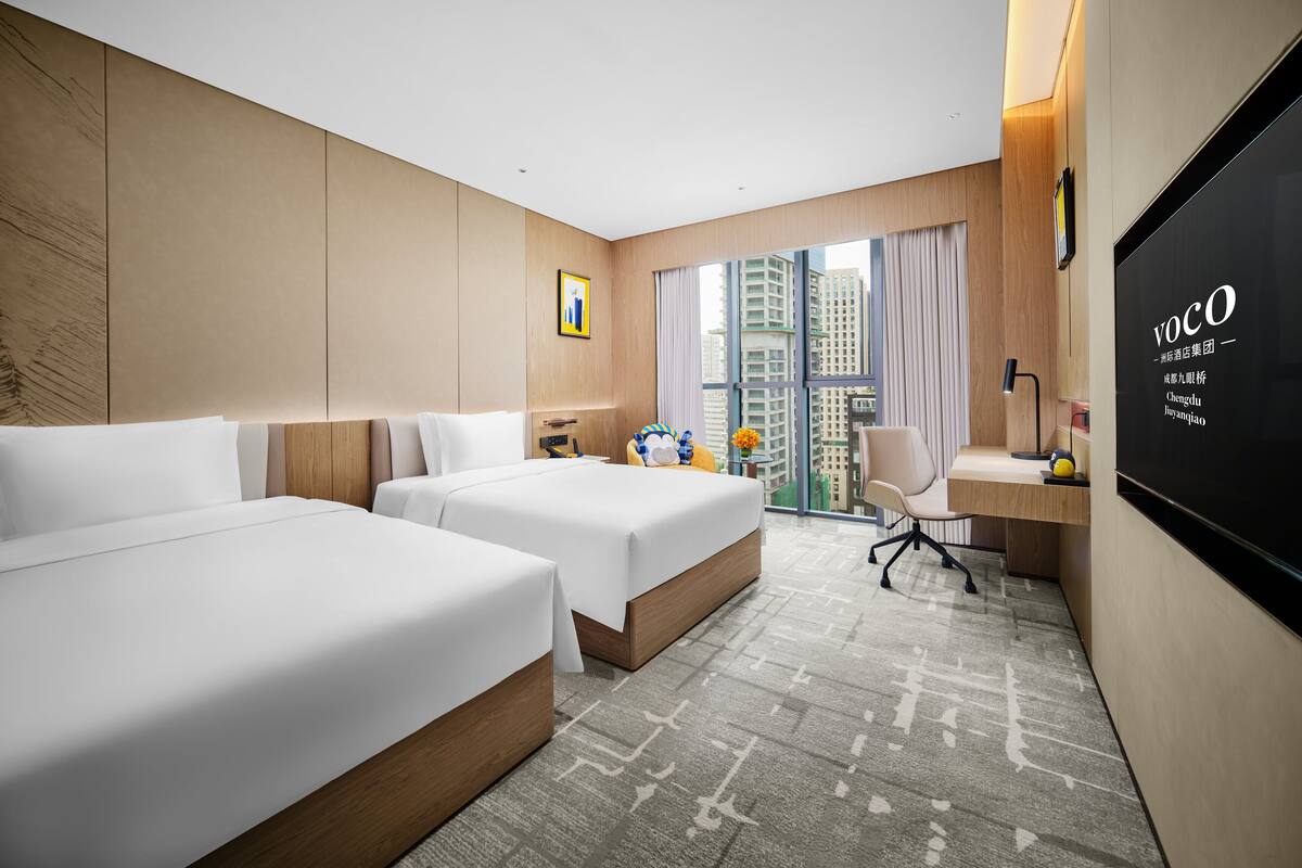 A hotel room with two beds, a desk, a chair, a TV, and a large window with a city view.