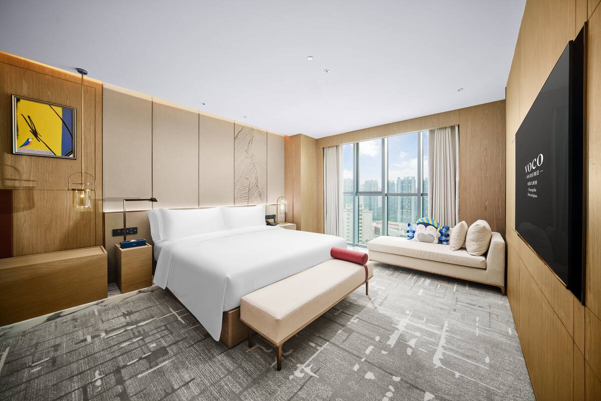 A modern hotel room with a large bed, a bench, a sofa, a TV, and a city view.
