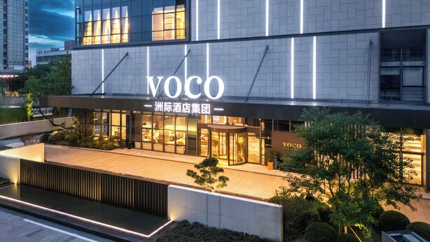Un hôtel moderne, reconnaissable à son grand panneau « VOCO », doté d’une façade en verre et d’une entrée bien éclairée.