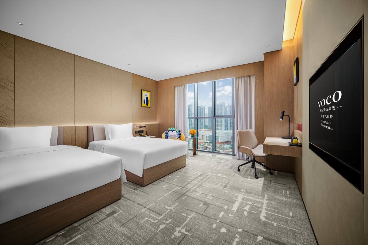 A hotel room with two beds, a desk, a chair, a TV, and a large window with a city view.