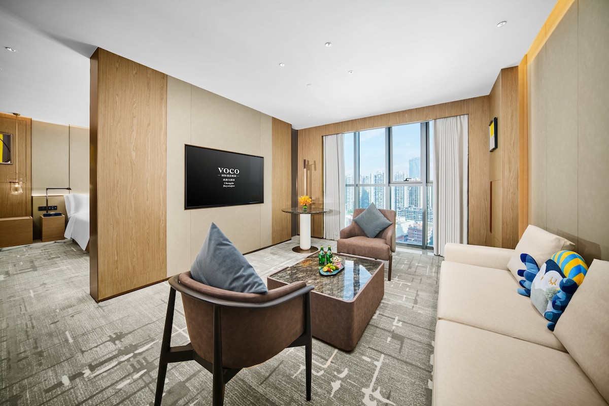 Habitación de hotel moderna con televisor de pantalla plana, sofá, mesa de centro con frutas y vistas a la ciudad.