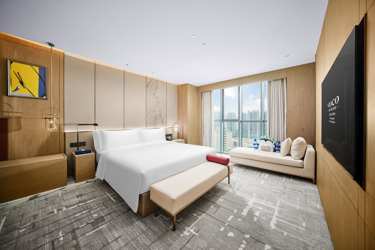 Una camera d'albergo moderna con un letto grande, una panca, un divano, una TV e vista sulla città.