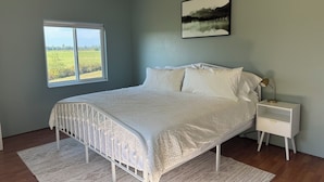 A bedroom with a bed, a nightstand, a lamp, a framed picture, and a window with a view of a field. - Charming 3-bedroom farmhouse in Astoria (Astoria)