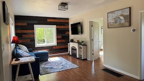 A modern living room with a wooden accent wall, a fireplace, a TV mounted above it, and a large window providing natural light. - Charming 3-bedroom farmhouse in Astoria (Astoria)
