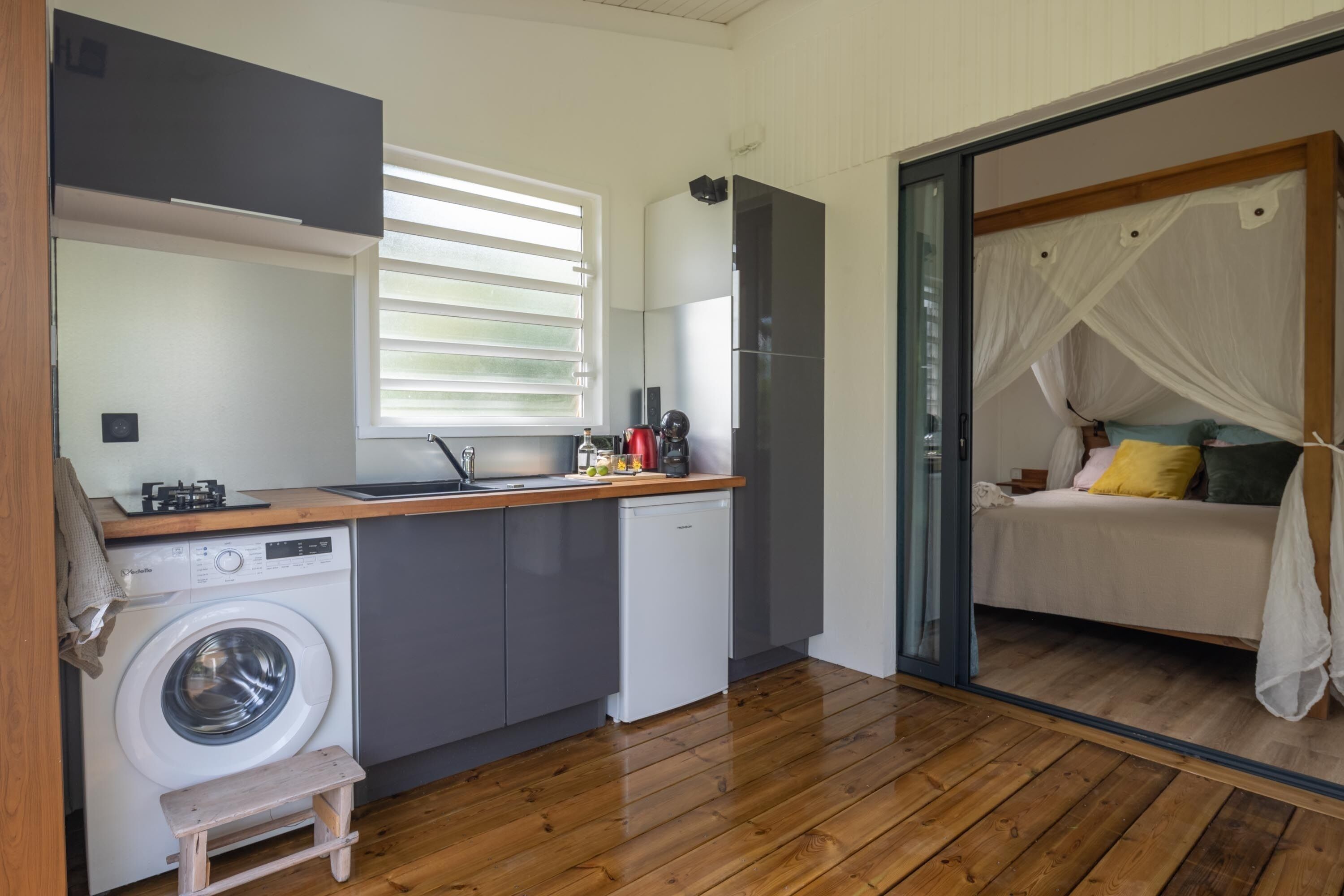 A compact kitchen with a washing machine, a small wooden stool, and a bed with a canopy in the adjacent room.