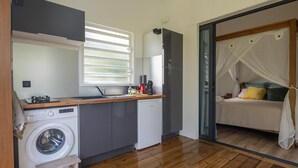 A compact kitchen with a washing machine, a small wooden stool, and a bed with a canopy in the adjacent room.