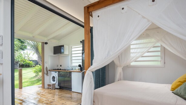 A bedroom with a canopy bed, a washing machine, and a kitchen area visible through an open door.