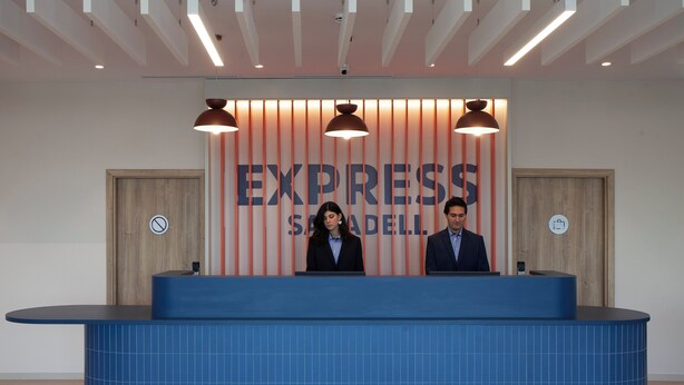 A reception desk with a blue tiled front, two attendants behind it, and a sign reading "EXPRESS SADELLI". - Holiday Inn Express & Suites Barcelona Sabadell by IHG (Sabadell)