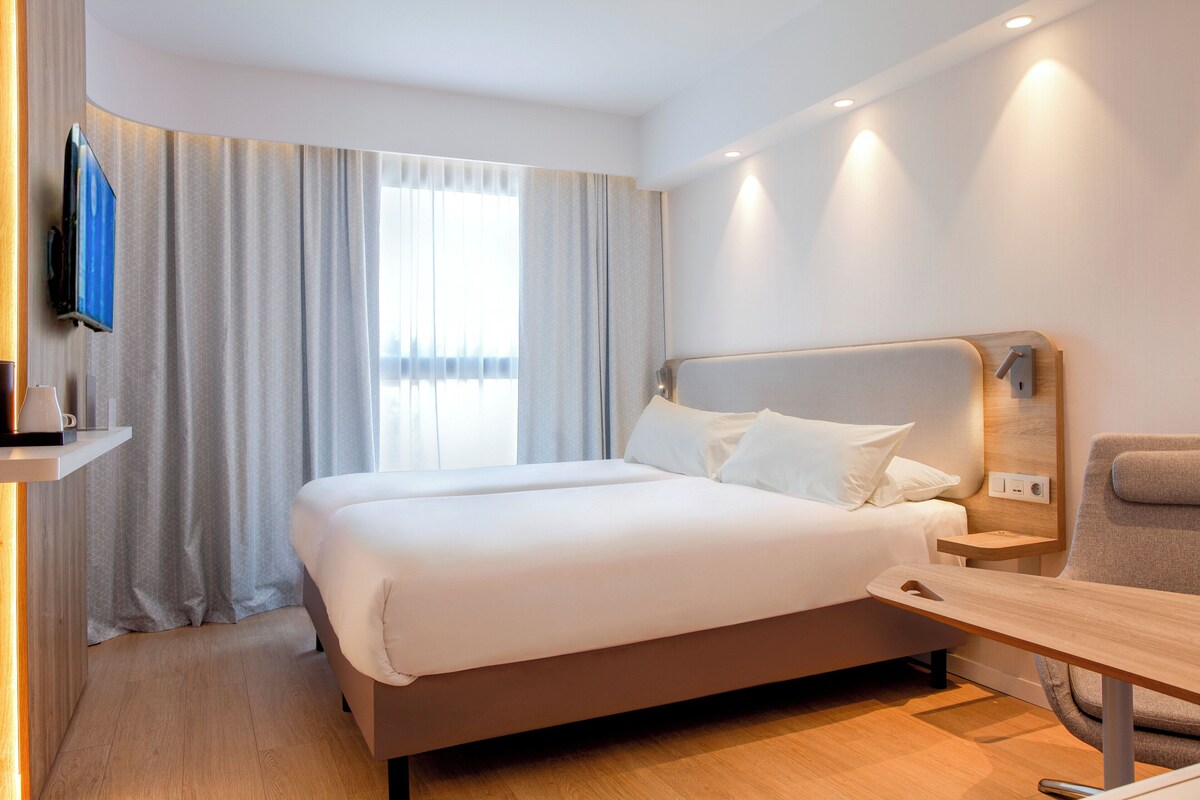 A modern hotel room with a large bed, a bedside table, a chair, a TV mounted on the wall, and a window with curtains.