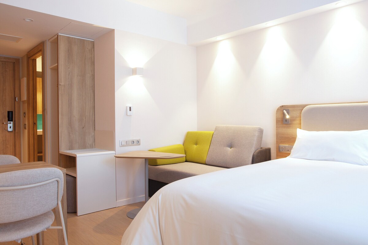 A modern hotel room with a bed, a desk, and a seating area.