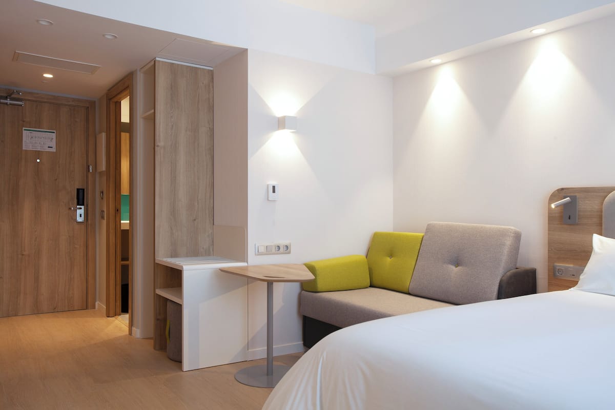 A modern hotel room with a bed, a desk, and a seating area with cushions.