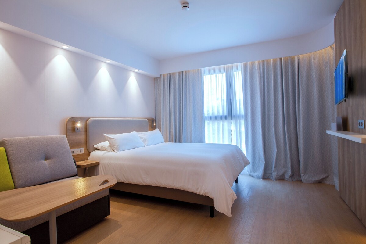 A modern hotel room with a bed, a seating area, a flat-screen TV, and a large window with curtains.