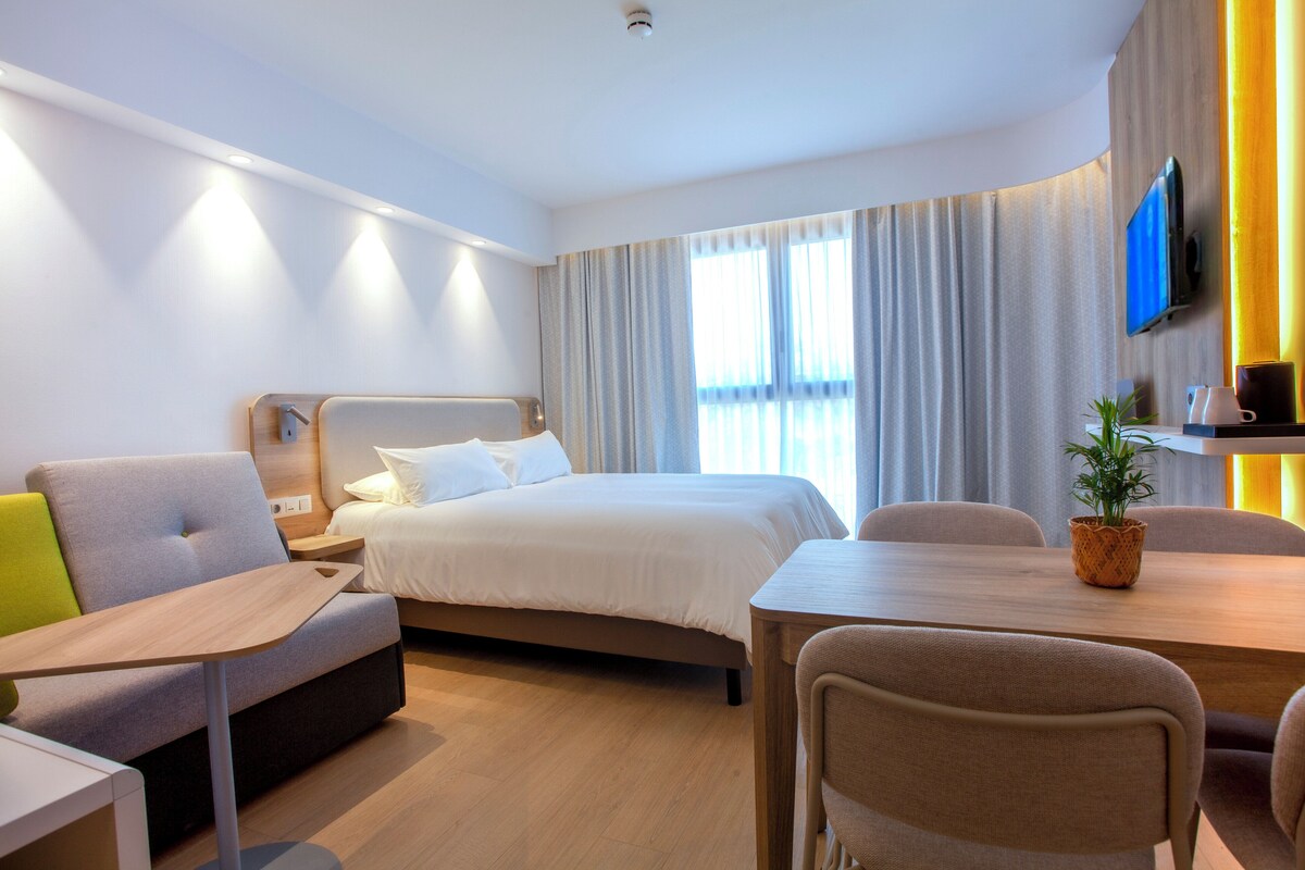 A modern hotel room with a bed, a sofa, a dining table, and a TV mounted on the wall.