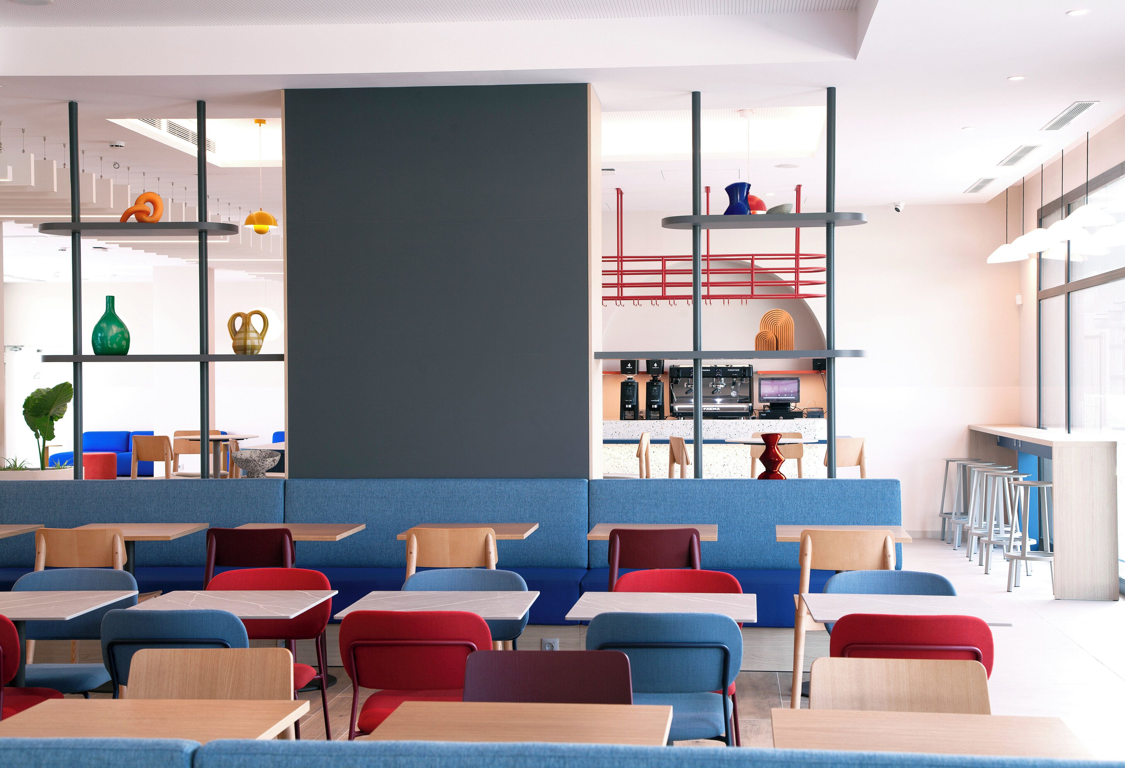 A modern cafeteria with blue and red seating, a central serving area, and a large window providing natural light.