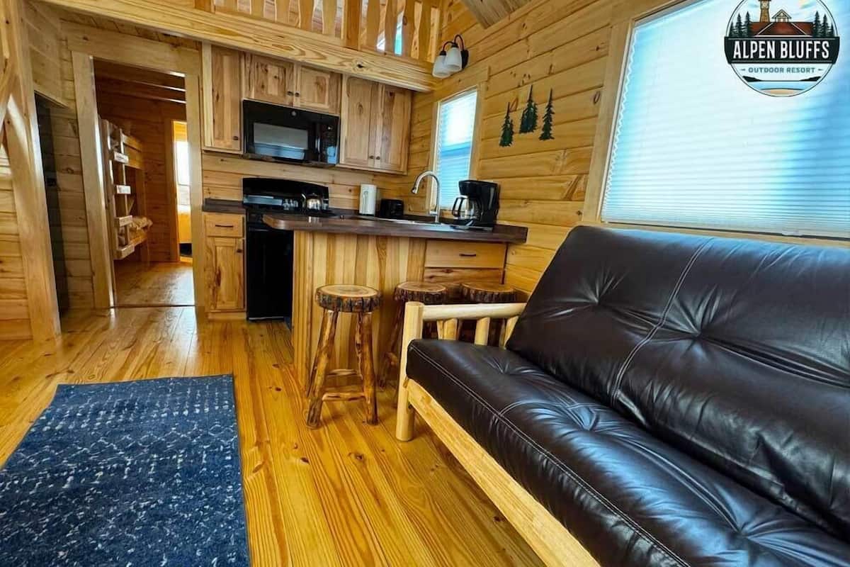 A cozy cabin interior with a wooden interior, a black leather sofa, a kitchen area, and a dining table with stools.