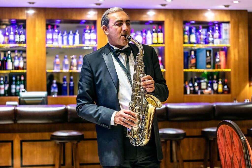 Une personne joue du saxophone dans un bar, avec des étagères remplies de bouteilles en arrière-plan.