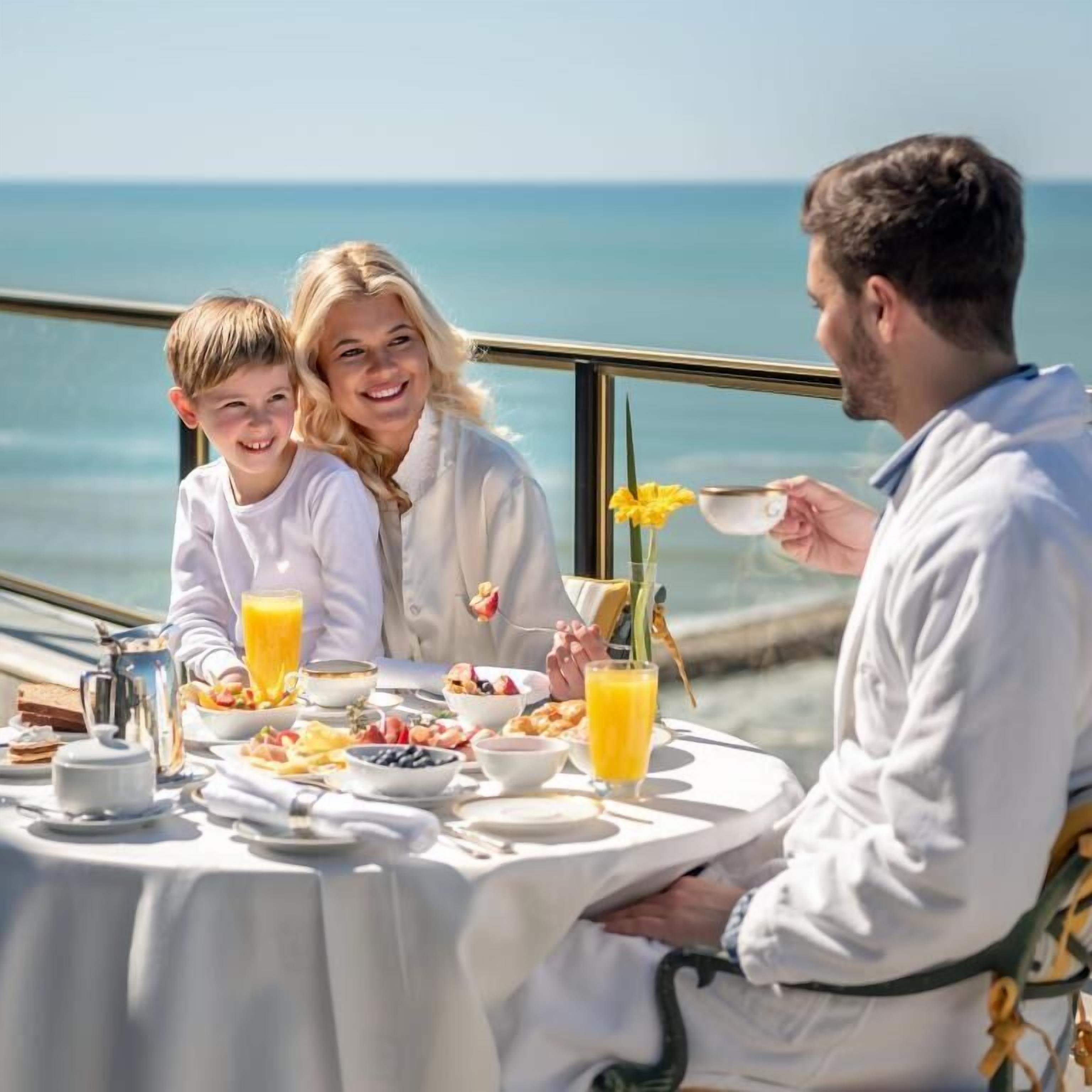 Une table à manger avec des aliments pour le petit-déjeuner, une vue sur la mer et une personne qui sert les plats.