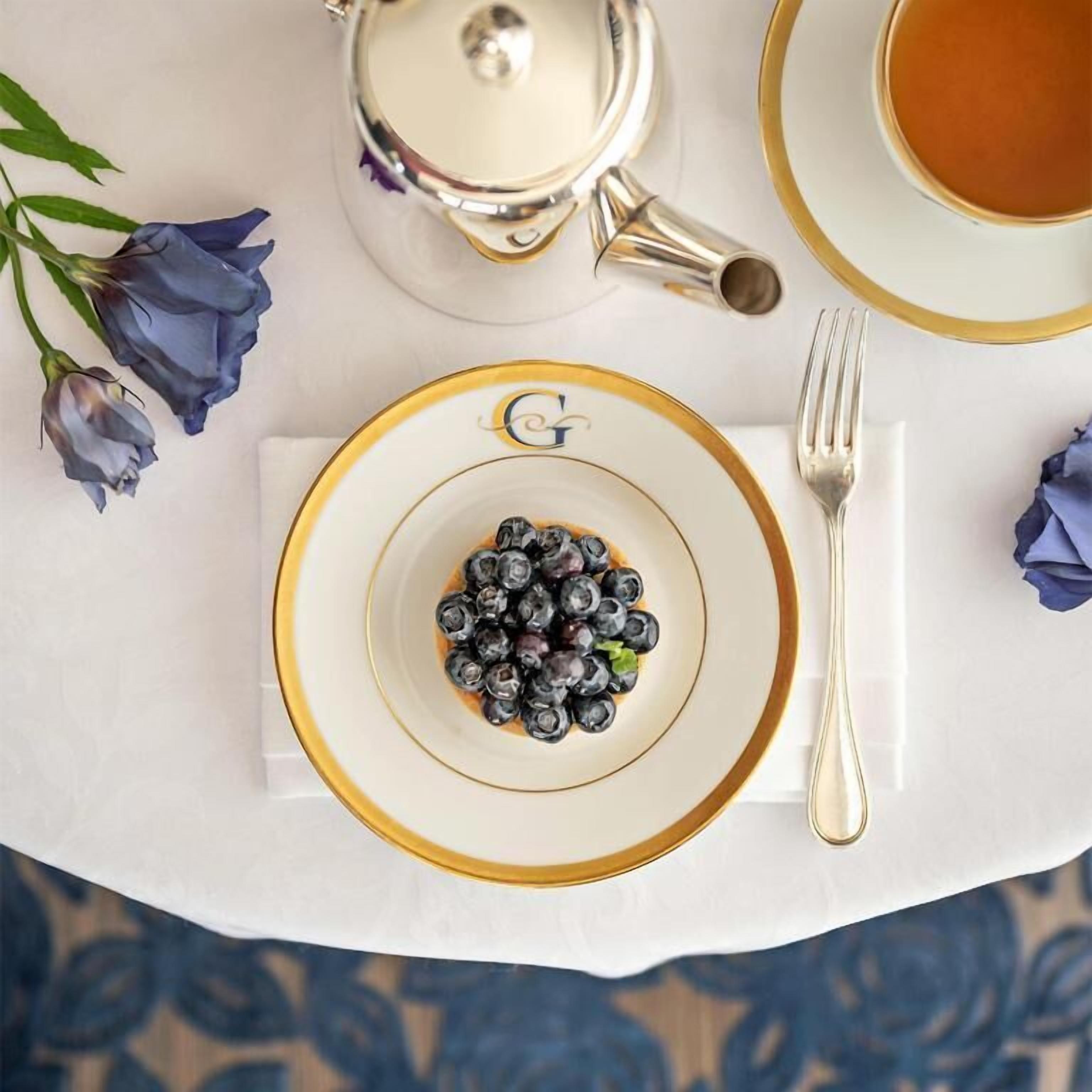 Ein Tisch ist mit einem Tee-Service, einer Tasse Tee, einer Schale mit Blaubeeren und einer Gabel auf einer weißen Tischdecke gedeckt.