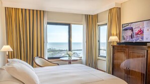 Une chambre d’hôtel avec un lit, une chaise, une petite table avec un vase, une télévision et une vue sur la mer.